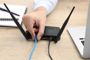 Man inserting cable into Wi-Fi router at wooden table indoors, closeup