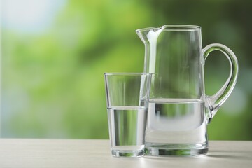Jug and glass with clear water on white table against blurred green background, closeup. Space for text