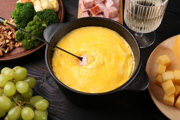 Fondue pot with melted cheese, fork and different products on black wooden table