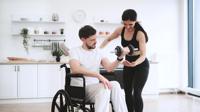 Rehabilitation of disabled people concept. Young physiotherapist or wife in sportswear helping Caucasian male patient in wheelchair exercise at home. Handicapped mature man training with dumbbells.