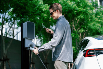 Young man travel with EV electric car charging in green sustainable city outdoor garden in summer...
