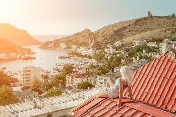 Woman sits on rooftop, enjoys town view and sea mountains. Peaceful rooftop relaxation. Below her,...
