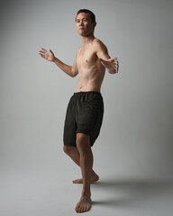Fototapeta premium full length portrait of fit asian male model, shirtless with muscles. gestural ti chi inspired posing with arms reaching out. Isolated on a white studio background with moody silhouette.