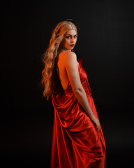 Fototapeta premium Close up portrait of beautiful blonde model wearing flowing red silk toga gown and crown, dressed as ancient mythological fantasy goddess. Graceful elegant pose isolated on dark studio background.