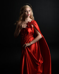 Fototapeta premium Close up portrait of beautiful blonde model wearing flowing red silk toga gown and crown, dressed as ancient mythological fantasy goddess. Graceful elegant pose isolated on dark studio background.