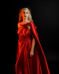 Naklejka premium Close up portrait of beautiful blonde model wearing flowing red silk toga gown and crown, dressed as ancient mythological fantasy goddess. Graceful elegant pose isolated on dark studio background.