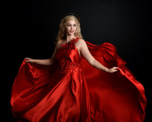 Close up portrait of beautiful blonde model wearing flowing red silk toga gown and crown, dressed as ancient mythological fantasy goddess. Graceful elegant pose isolated on dark studio background.