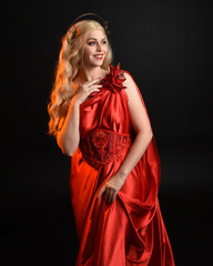 Close up portrait of beautiful blonde model wearing flowing red silk toga gown and crown, dressed as ancient mythological fantasy goddess. Graceful elegant pose isolated on dark studio background.