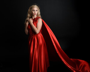 Fototapeta premium Close up portrait of beautiful blonde model wearing flowing red silk toga gown and crown, dressed as ancient mythological fantasy goddess. Graceful elegant pose isolated on dark studio background.