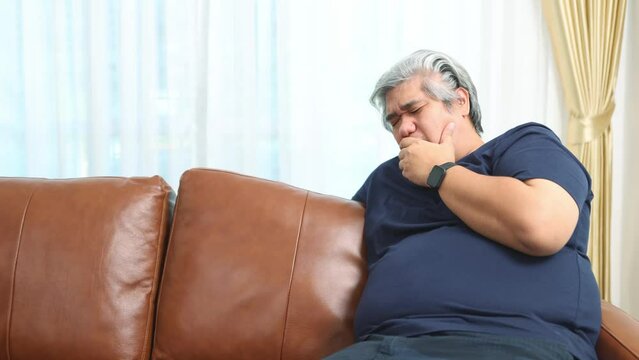 Obese asian man sit on the sofa has health problems suffering from respiratory and lung diseases chronic cough severe chronic bronchitis lung infection symptoms choking on food or saliva.