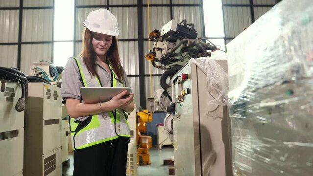 Female engineer inspects control cabinet digital tablet checks electrical switch buttons controls industrial robot in warehouse covered clear plastic dustproof and maintenance waiting electrical test.