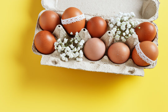 Easter eggs decorated with lace and white spring flowers in carton box on yellow background. Easter holiday, aesthetic top view of festive chicken egg in container, celebration life still