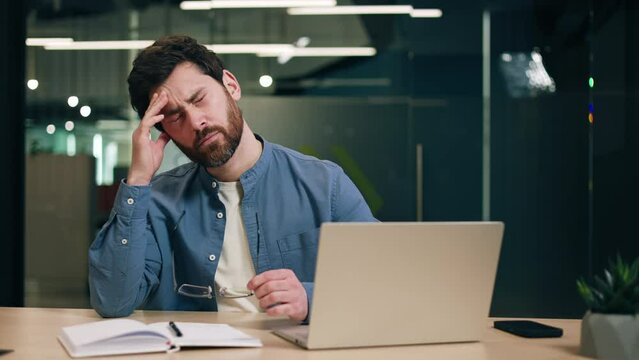 Stressed businessman sitting by table with computer and daybook and touching forehead with closed eyes. Hardworking startup runner trying meeting deadlines for making profitable deals with clients.