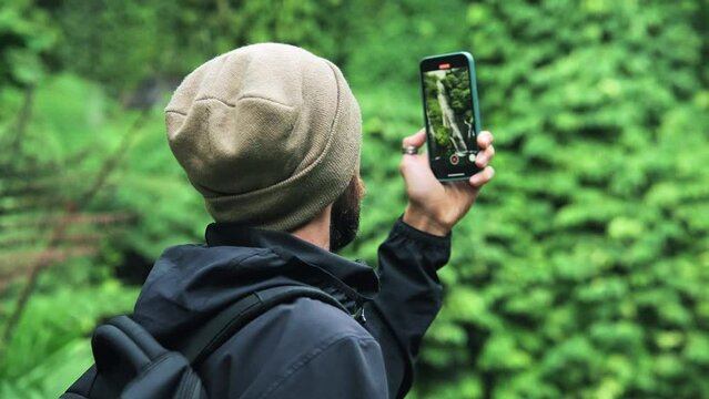 Adult Man Travels in Green Nature Taking Phone Photo of Forest Waterfall. Concept of Young Touring Person at Trip Destination of Jungle Water Fall. Caucasian Traveler Makes Videos on Journey Lifestyle