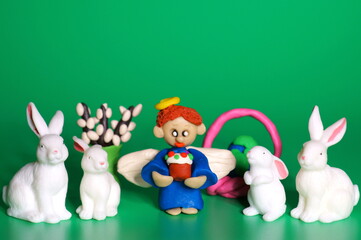 An angel figurine with an Easter cake on a green background. Nearby are Easter hares, willows and an Easter egg in the form of the planet earth.