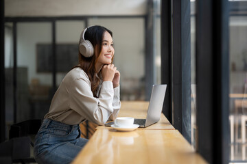 A young woman enjoys music wearing headphones. Looking at a laptop in a bright coffee shop With a cup of coffee next to her