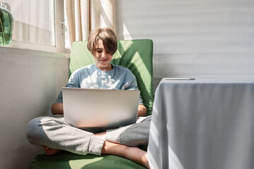 Teenager boy is sitting and holding laptop computer on knees, typing, communicating online, looking...