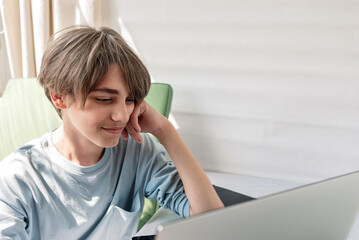 Portrait of teenage boy, sitting indoor and looking on laptop screen, lifestyle, bright natural sunlight. Learning or communication online at home, video call or meeting