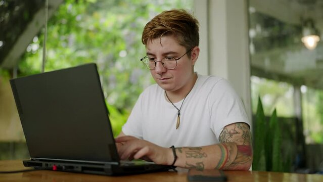 Focused tattooed transgender person types on laptop in sunlit modern cafe boasting expansive glass walls and lush green setting, embodying diverse remote work lifestyle. Slow motion.