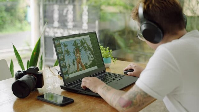 Transgender photo editor works on retouching images at laptop in coworking space. Tattooed creative with headphones revises photos, celebrates diversity, embodies digital nomad lifestyle. Slow motion.