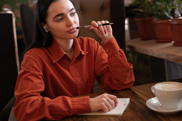 Pensive young female writer with notepad and pen looking away and thinking while working on new article sitting in modern cafe