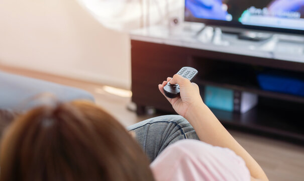 Woman relaxing on the couch, she is using the remote control and choosing a TV show or movie on the television menu.