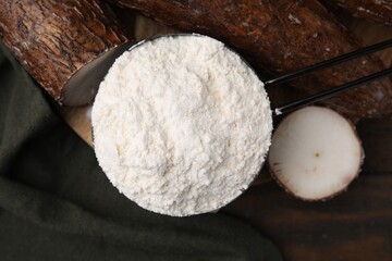 Scoop with cassava flour and root on wooden table, top view