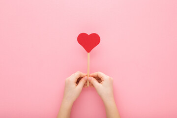 Little child hands holding red paper heart shape on wooden stick on light pink table background....