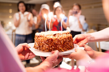 Hands of young people holding a happy birthday cake with their friends Candle flame lights on birthday cake during celebration - obrazy, fototapety, plakaty