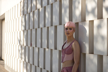 Young, attractive, gay man, heavily makeup, with pink hair and top, posing looking at the camera, relaxed and calm, leaning against a wall of white blocks. LGTBIQ+ concept, gay, pride, makeup.