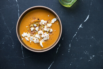 Bowl of sweet potato cream-soup topped with feta and pistachios, flat lay on a black marble background, horizontal shot with space