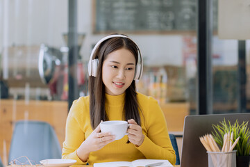 Happy asian young businesswoman in wireless headphones and drinking a coffee sitting at desk table working on laptop in cafe. 