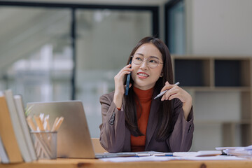 Happy asian young businesswoman using digital smartphone sitting in office working space, Asian female employee using laptop talking on the phone at workplace.