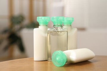 Mini bottles of cosmetic products and green branch on wooden table against blurred background, closeup