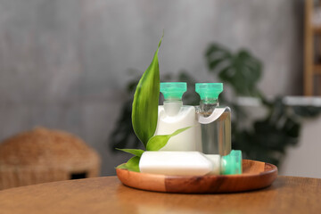 Mini bottles of cosmetic products and green branch on wooden table against blurred background. Space for text