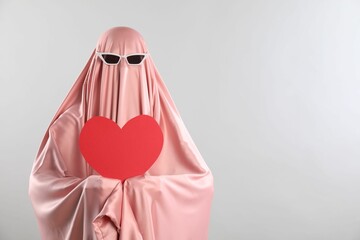 Cute ghost. Woman in pink sheet with sunglasses and red heart on light grey background, space for...