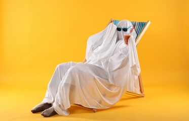 Fototapeta na wymiar Person in ghost costume and sunglasses with glass of drink relaxing on deckchair against yellow background