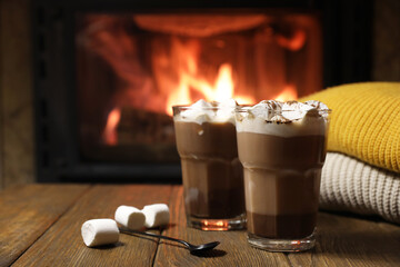 Glasses with hot cocoa, marshmallows and sweaters on wooden table near fireplace, space for text