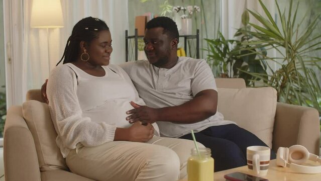 Medium long of African pregnant wife and happy husband resting on sofa and talking together at home