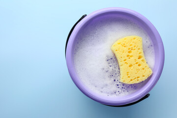 Bucket with foam and sponge on light blue background, top view. Space for text