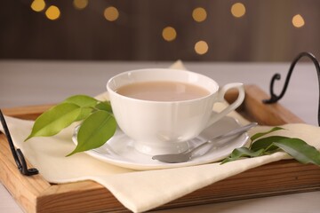 Cup of aromatic tea with milk, spoon, saucer and green leaves on table