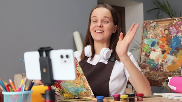 Joyful Caucasian brown haired woman recording master class of drawing with smartphone on tripod broadcasting livestream wearing apron painting at her workplace in art studio