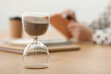 Hourglass with flowing sand on desk. Woman working indoors, selective focus