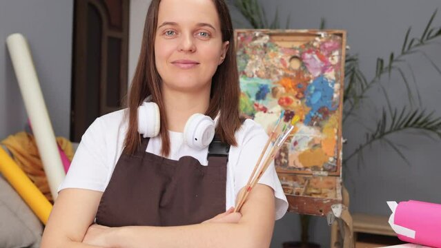 Smiling cheerful Caucasian brown haired woman wearing apron sitting at workplace in drawing studio enjoying her hobby creating works of art. Imaginative art exploration.