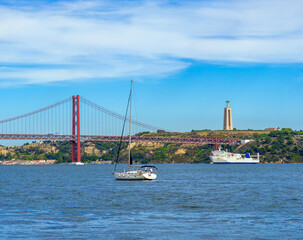 Sailboat and white ferry sailing along the Tajo River with the 25 de Abril steel suspension bridge...
