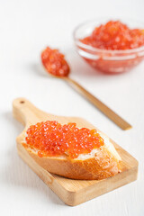 Homemade sliced loaf of bread or canape with butter and salty natural salmon fish roe or red caviar served on chopping board on white wooden table with spoon and glass bowl as celebration appetizer