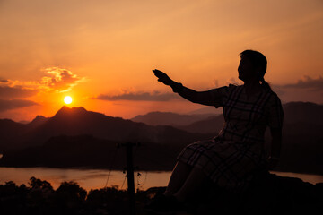 Fototapeta premium Ombre d'une femme asiatique assise tendant la main vers le coucher de soleil au-dessus d'une montagne