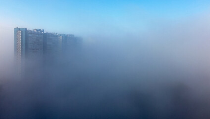 Fototapeta premium A white-and-blue multi-storey residential building is shrouded in fog in the early spring morning.