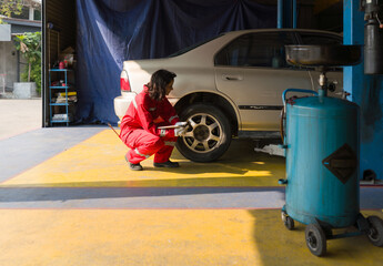 Young mechanic in a red coverall working on a car inside a workshop or garage. She is squatting beside the rear wheel of a car and inspecting, repairing, or performing maintenance on the wheel or tire