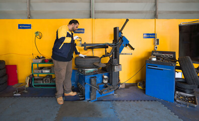 Fototapeta na wymiar A technician in uniform and glove at an automotive service center working with a tire-changing machine. There are various garage equipment and tools in the automotive maintenance and repair sector.